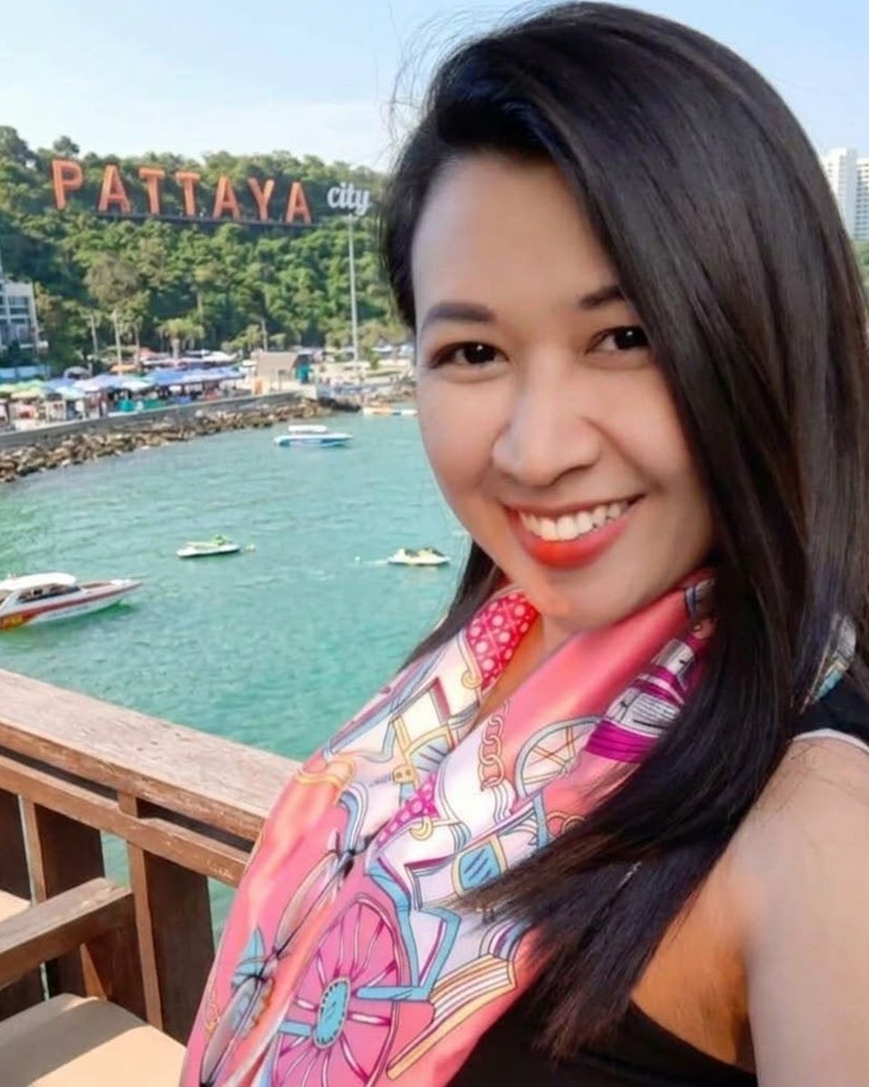 Danita, smiling, with long dark hair, a pink patterned silk scarf and a black top, on a pier above Pattaya Bay with the PATTAYA hillside sign behind her