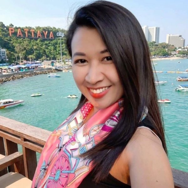 Danita, smiling, with long dark hair, a pink patterned silk scarf and a black top, on a pier above Pattaya Bay with the PATTAYA hillside sign behind her