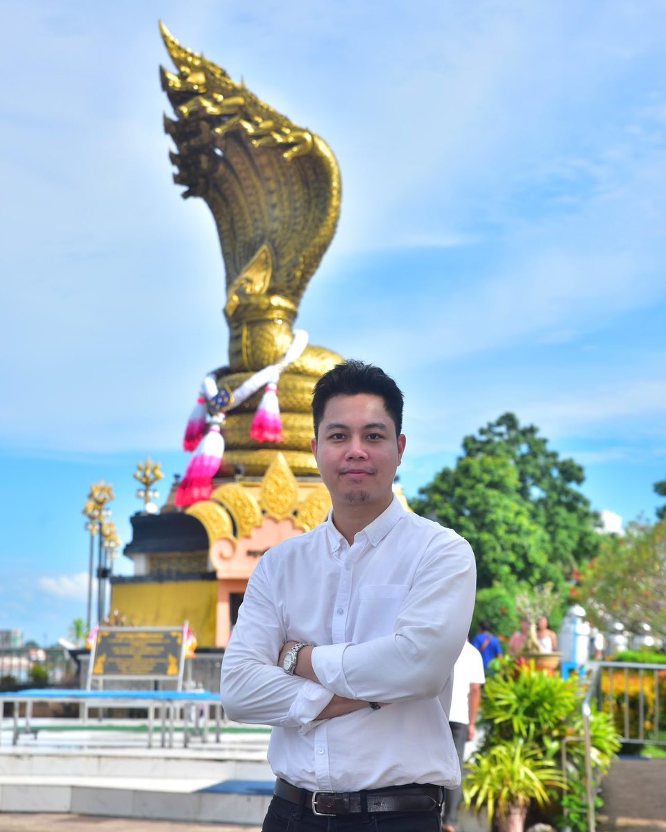 Thaksin (Taki) in a white shirt, arms folded, in front of a golden Naga statue under a blue sky