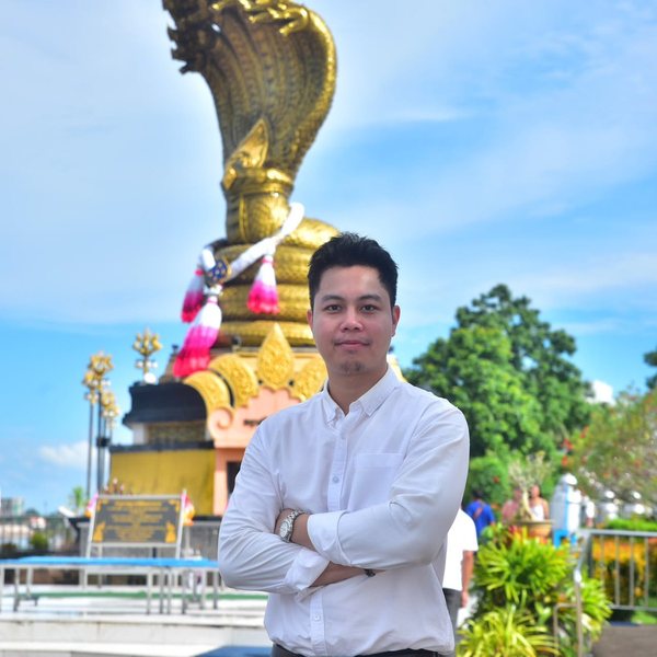 Thaksin (Taki) in a white shirt, arms folded, in front of a golden Naga statue under a blue sky